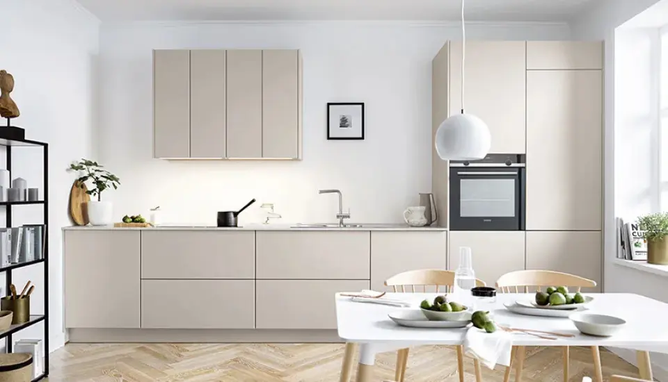 Helle, moderne Küche in sanftem Beige mit grifflosen Fronten, integriertem Backofen und schlanker Arbeitszeile; kombiniert mit weißem Esstisch, Holzstühlen, dekorierten Tellern und großen Fenstern für viel Tageslicht.